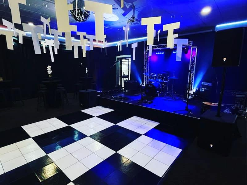 Image of an indoor music venue featuring a black and white checkered floor pattern, suspended abstract-shaped white lights, and a stage in the background with musical equipment illuminated by blue stage lights.